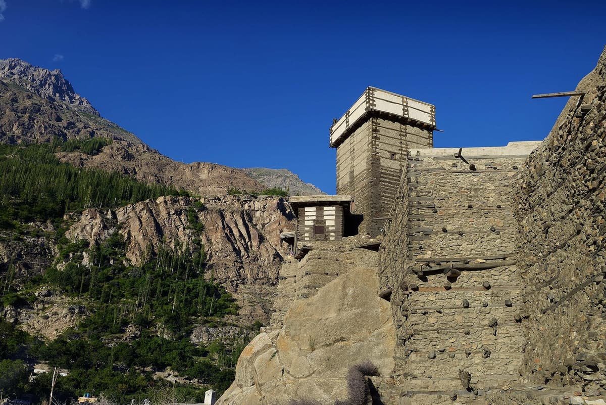 The stone tower of Altit Fort rises from a granite outcrop against a deep blue sky, beside poplar-covered mountain slopes.