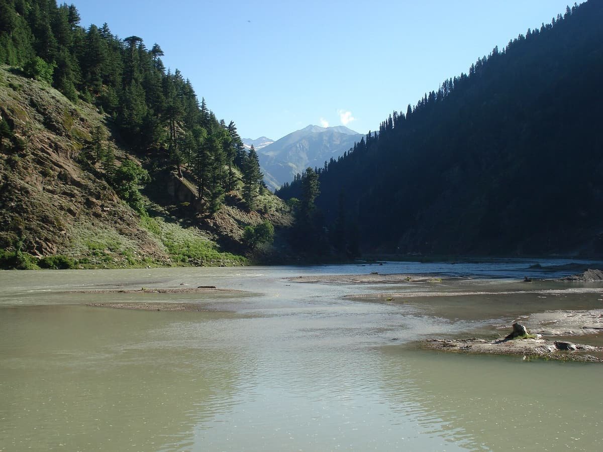 The broad Kunhar River flows past gravel bars between steep pine-covered slopes toward a distant peak.