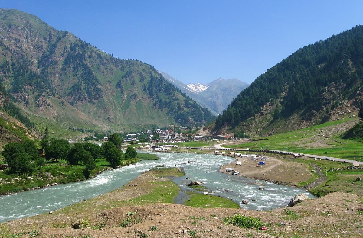 The Kunhar River curves through green meadows toward Naran town, set between steep forested mountains.