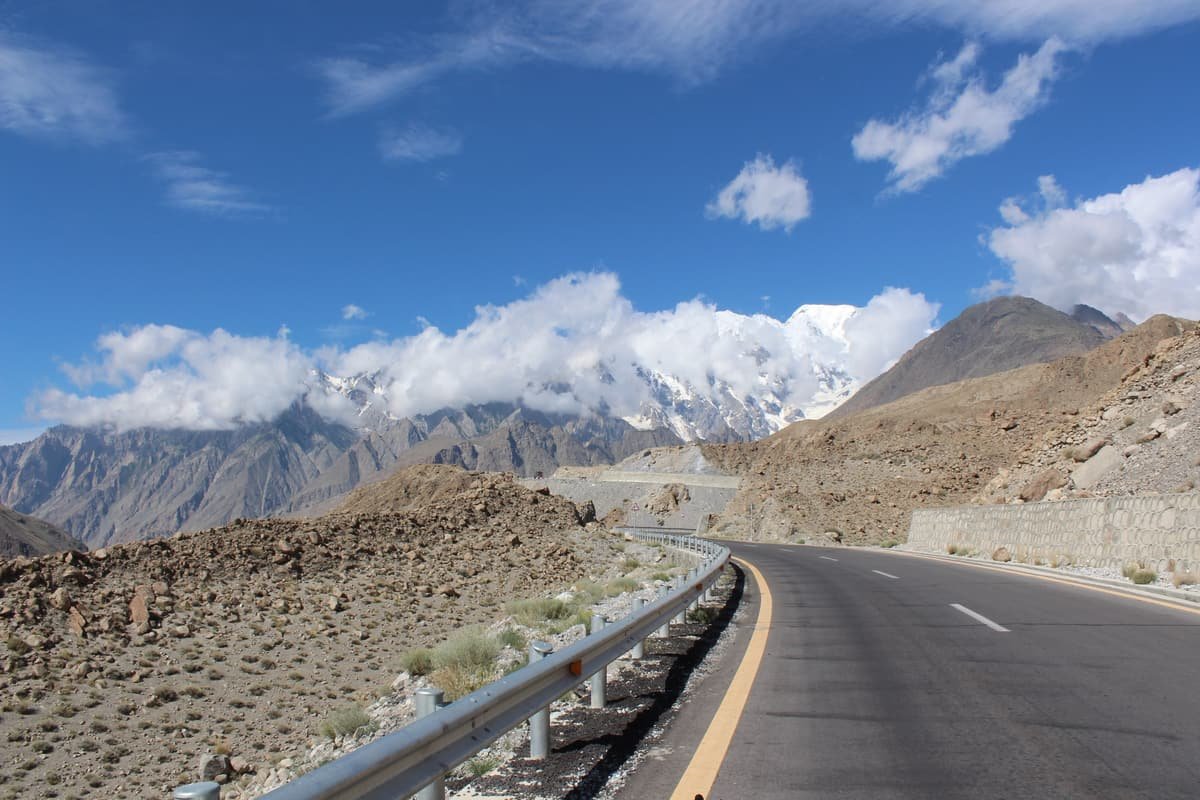 The Karakoram Highway curves past a guard rail and a stone wall towards snowy peaks half hidden in cloud