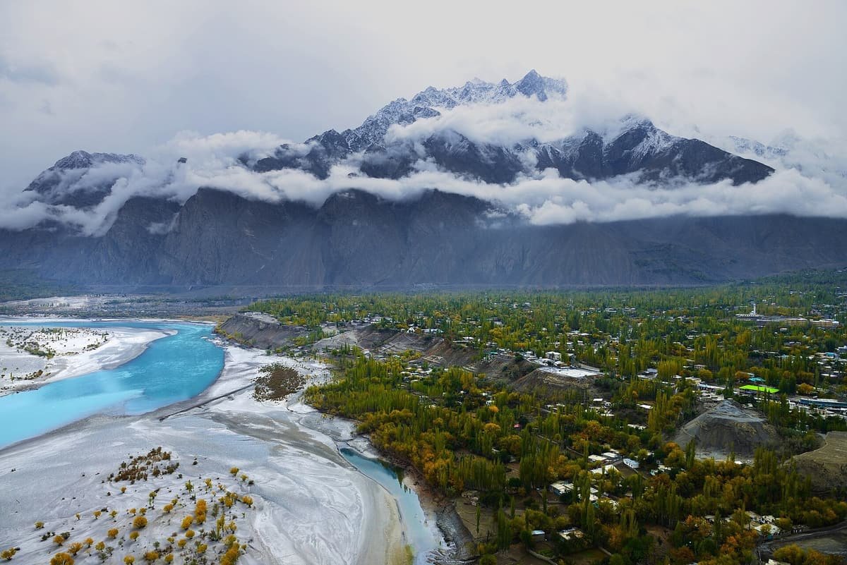 Skardu town sits among autumn trees beside the turquoise Indus River and its white sandbanks, below cloud-wrapped mountains.